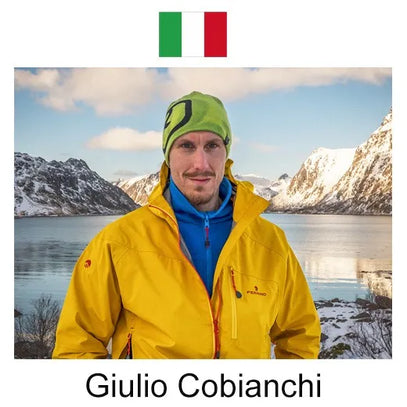 A person (in a bright yellow jacket, blue top, and green beanie) stands by a calm lake, with snow-capped mountains and a partly cloudy sky forming the scenic winter backdrop.