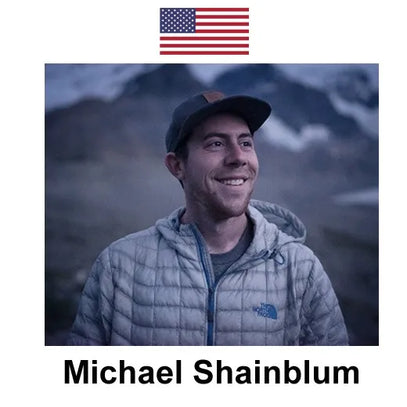 This image shows Michael Shainblum (in a gray The North Face puffer jacket and cap) smiling, with snow-dusted mountains behind him. An American flag sits above, and his name is labeled below.